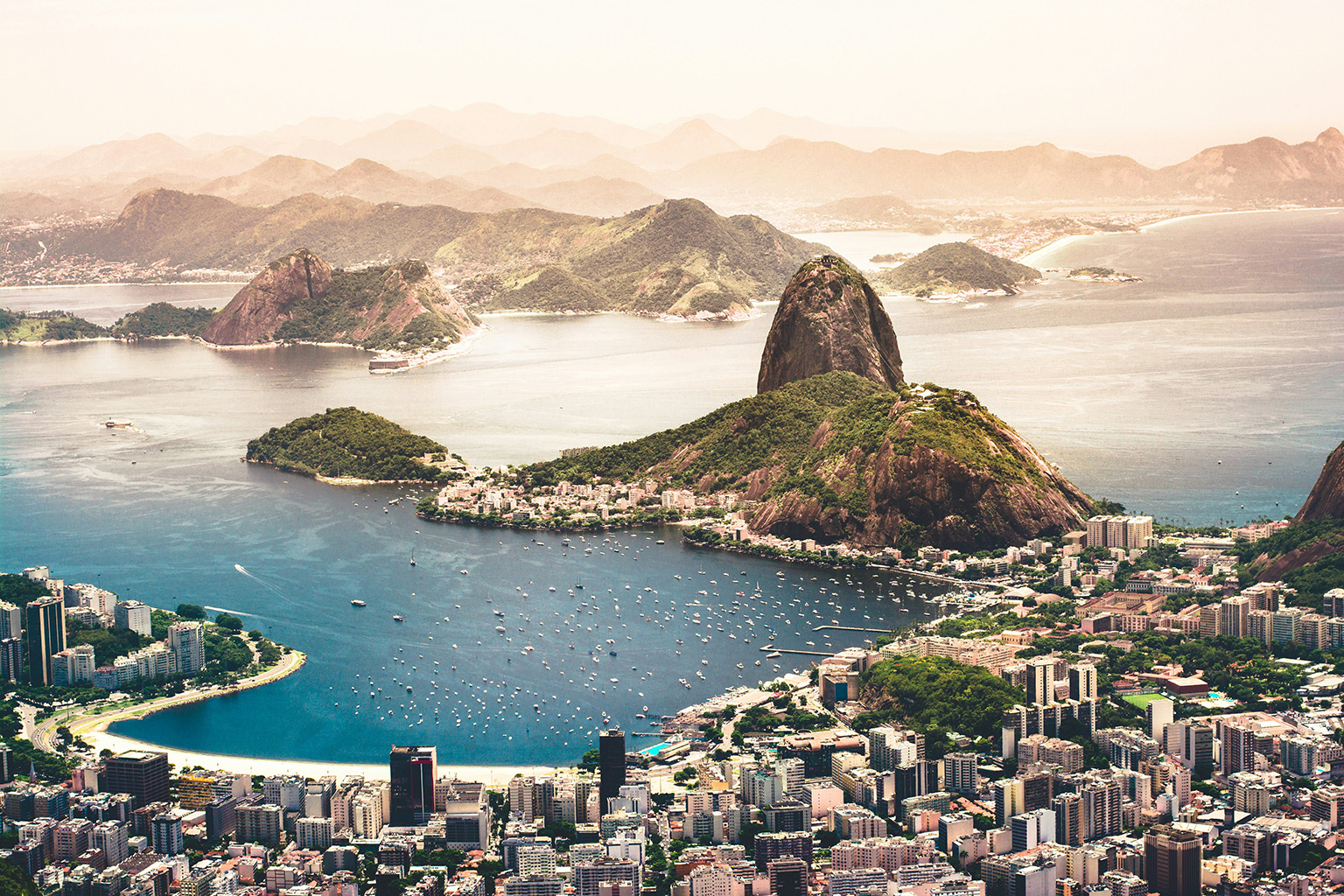 Sunset over the city of Rio de Janeiro, Brazil, with a view of the skyline, Sugarloaf Mountain, and Copacabana Beach illuminated by warm golden light.