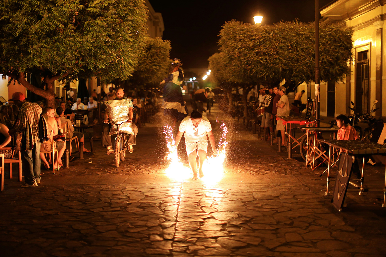 Night scene in a Latin American town with outdoor restaurants, people dining, and a fire performer in the street.