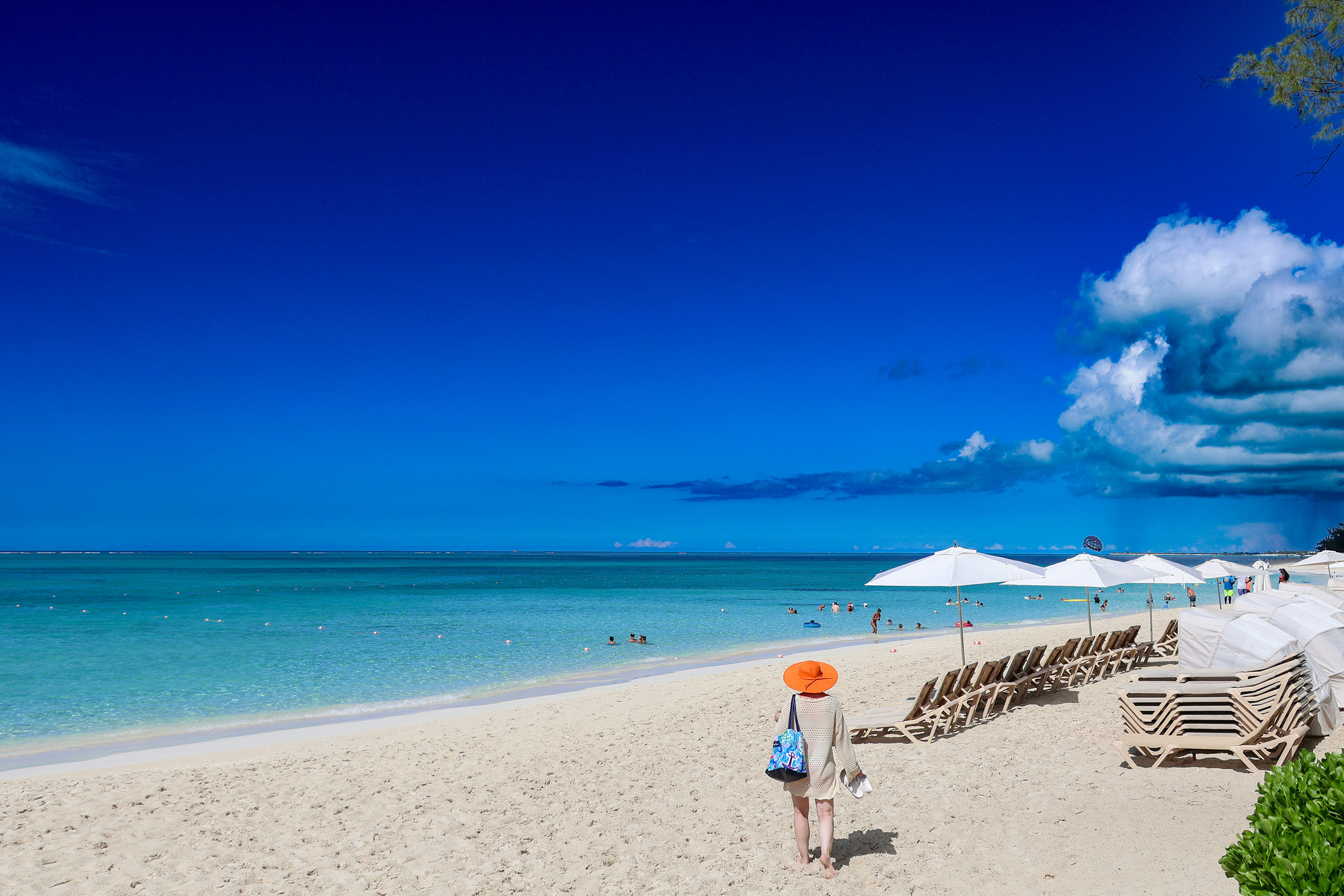 Beautiful Caribbean beach with blue sky, calm water, beach chairs, people swimming, and a woman walking towards the chairs.