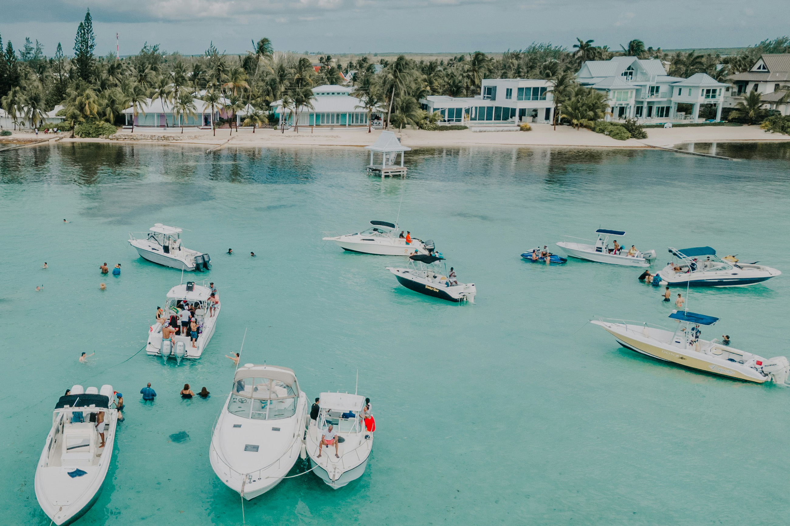 Typical life in the Caribbean, crystal-clear Caribbean water with moored boats, people swimming, and tropical houses along the shoreline.