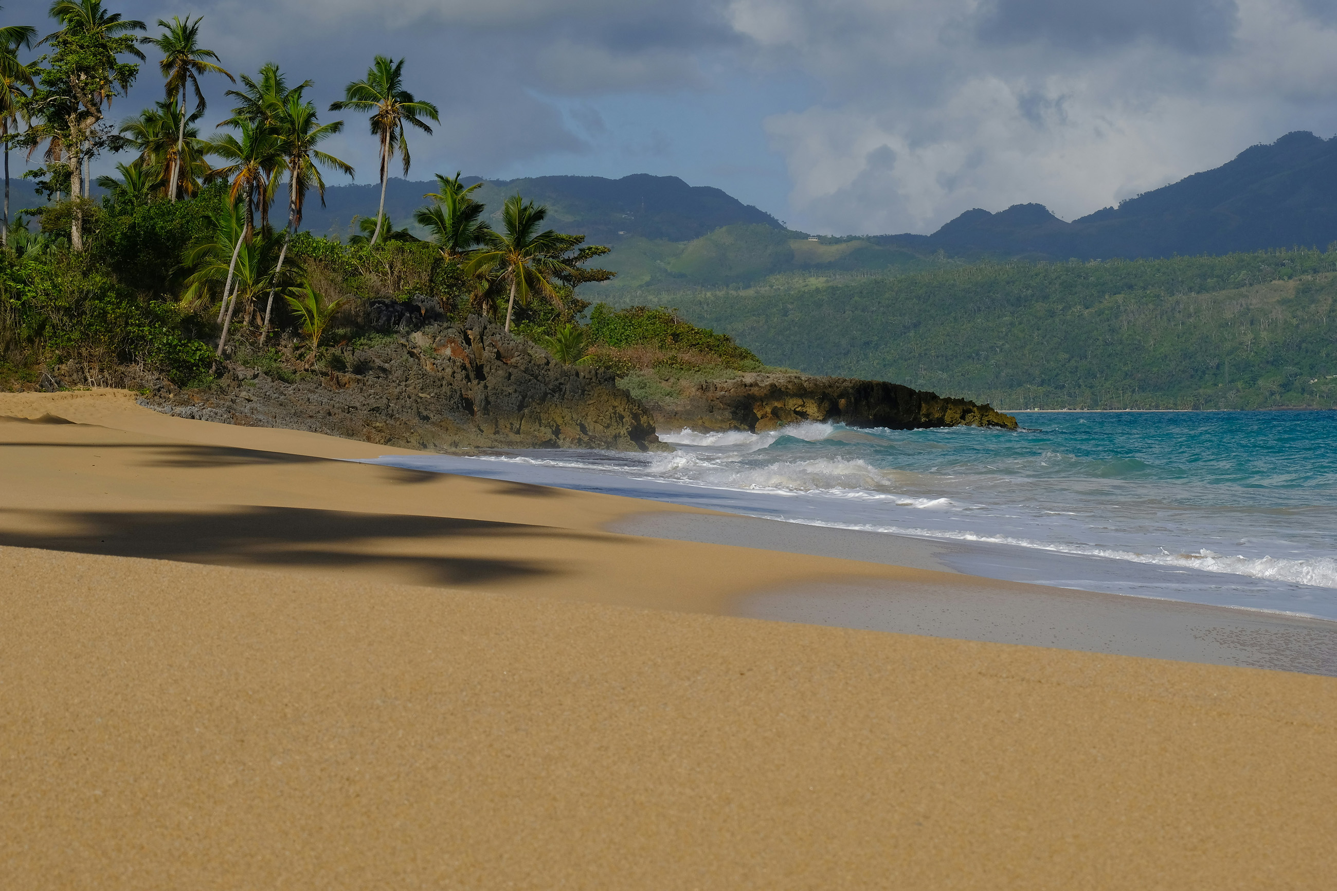Deserted tropical beach in the Caribbean with small waves, tropical forest, and mountains in the background.
