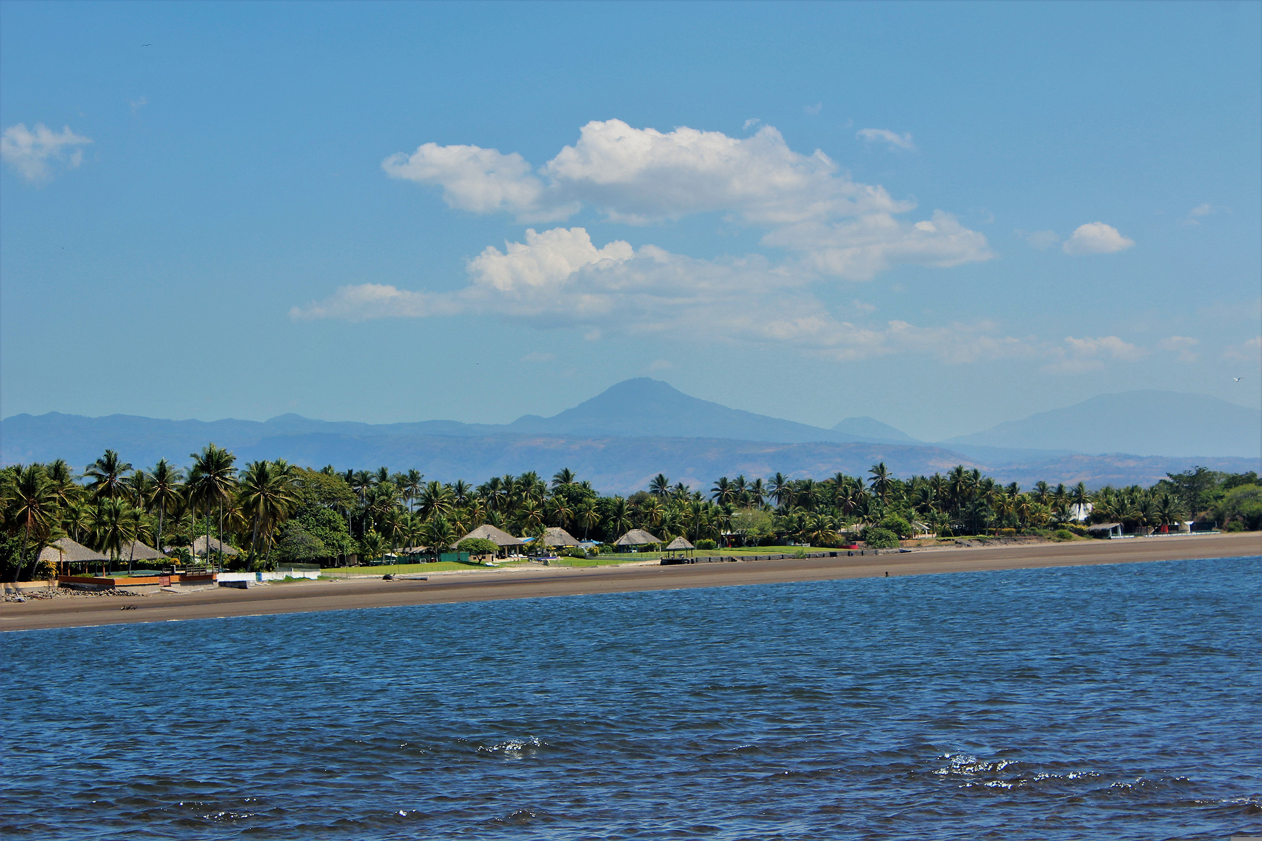 tree-lined-beach-ocena-El-Salvador Scenic beach in El Salvador with thatched-roof buildings, a tree line, and a volcano in the background, showcasing the beauty of the coastal landscape.