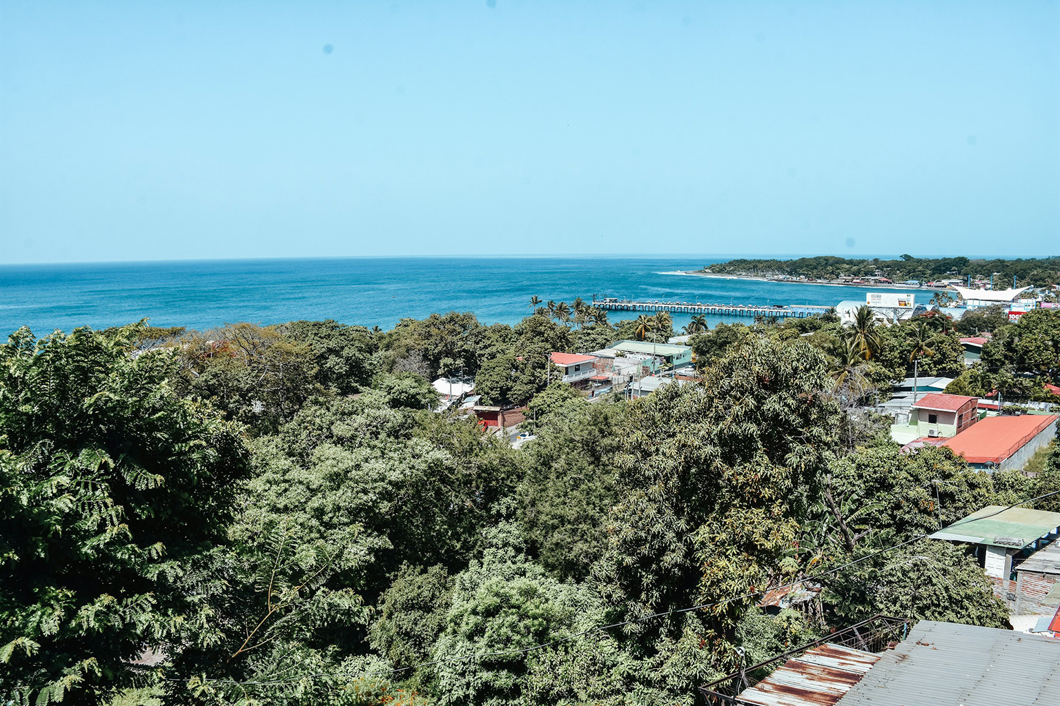 trees-house-roofs-ocean-El-Salvador Aerial view of lush greenery with house roofs and a small pier by the ocean, representing ideal places to live in El Salvador.