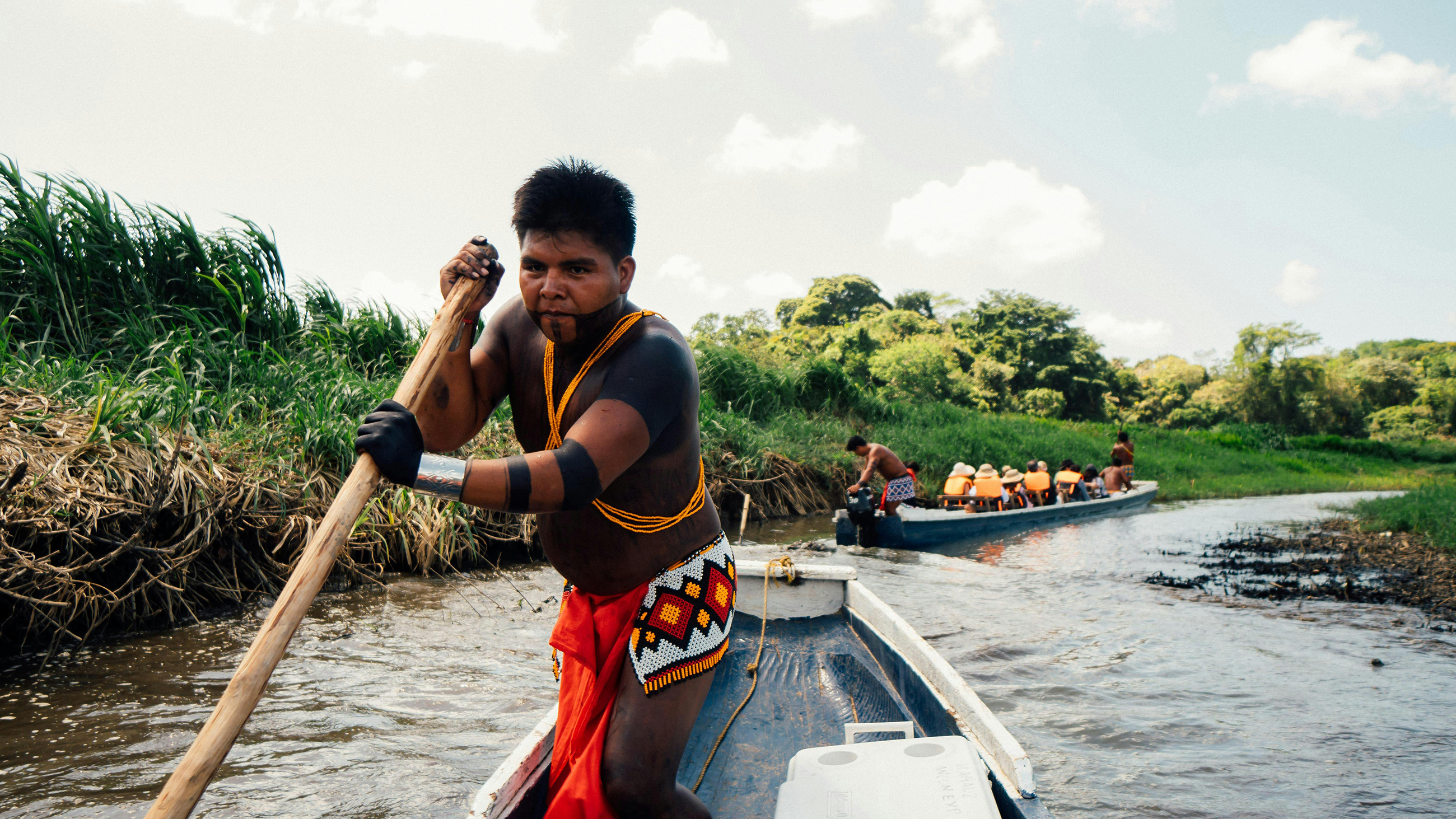 A local indigenous man in traditional dress paddling a boat along a river with tourists.