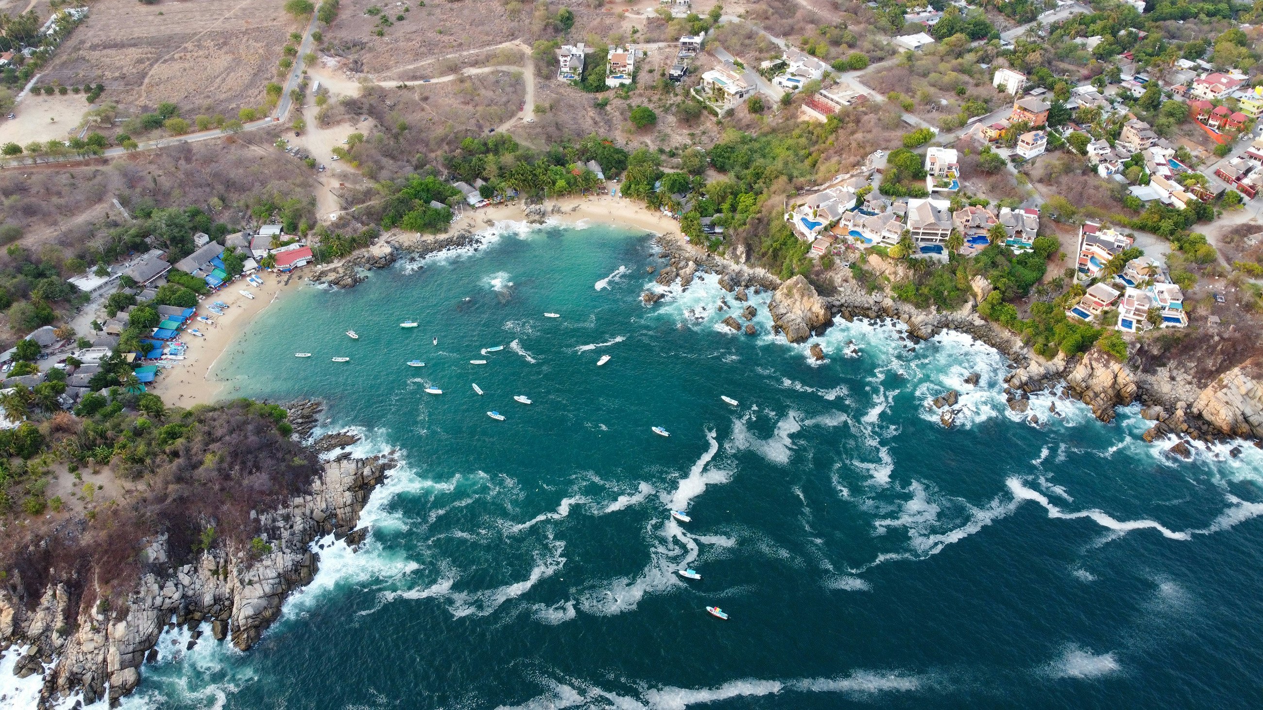 Aerial shot of Puerto Escondido’s rocky coastline, with homes along the cliffs and boats in the water