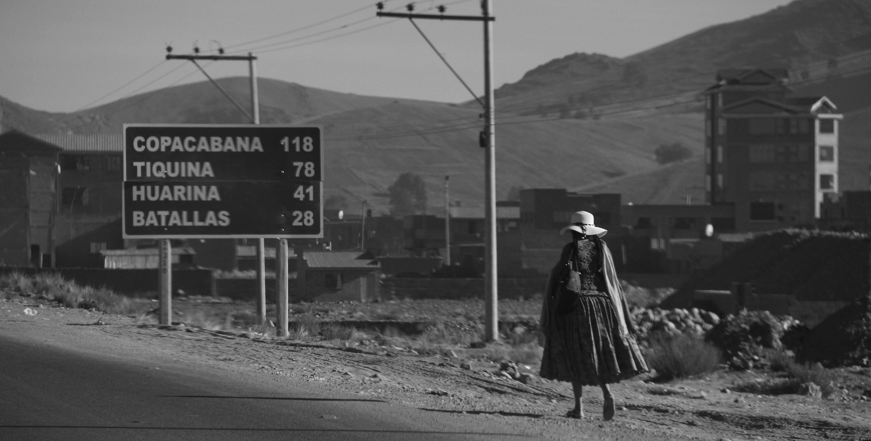 A woman in colorful traditional clothing walking along the highway with mountains and small buildings in the background, a common site when you live in Bolivia