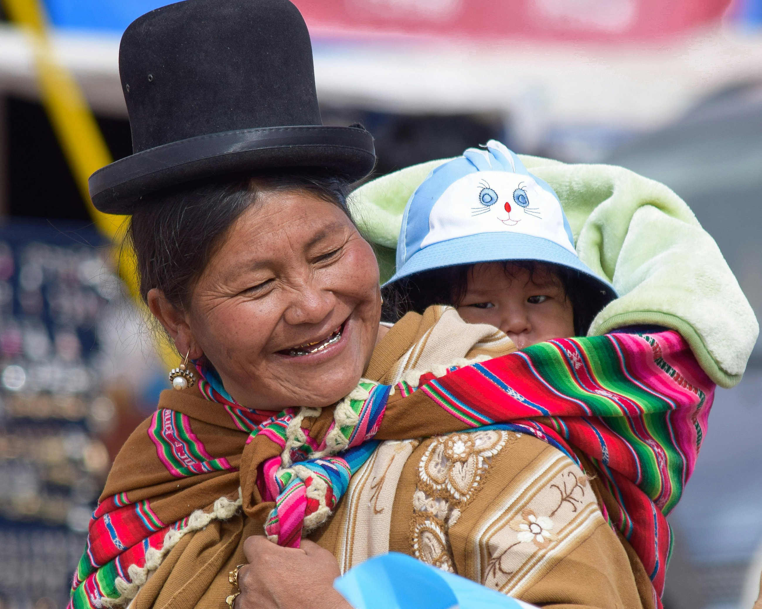 A close-up of a traditionally dressed Bolivian woman with a smile on her face, carrying her baby on her back.